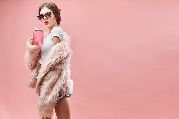 Garota com casaco de pele rosa faux shorts preto branco e óculos de sol contém bebida rosa com os olhos fechados ...