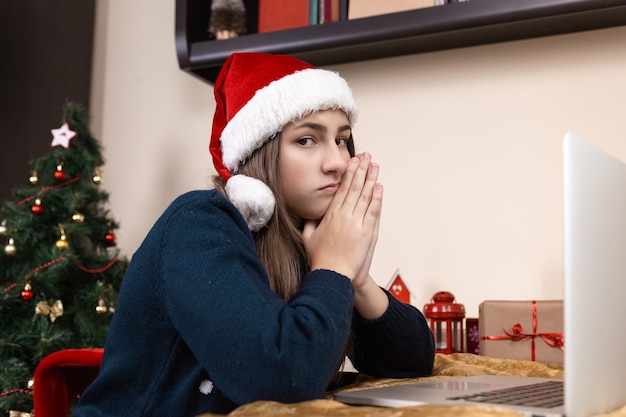 garota com chapéu de Papai Noel fala usando laptop para pais e amigos de videochamada. O quarto está decorado de forma festiva.