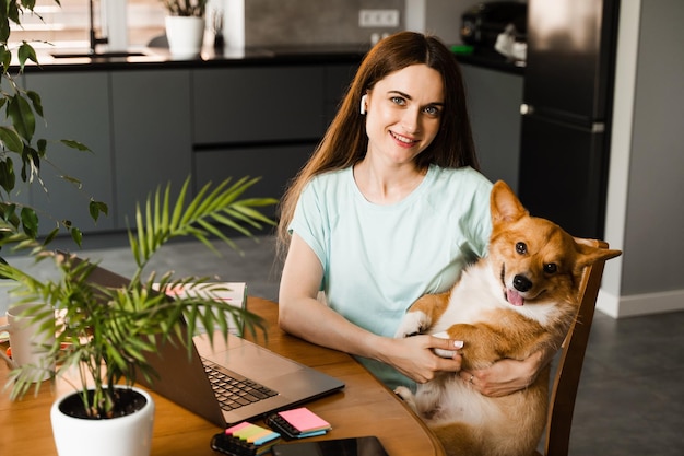 Garota com laptop sorri e brinca com cachorro Corgi em casa Mulher programadora trabalhando online e faz uma pausa para abraçar seu cachorro se divertindo com Welsh Corgi Pembroke