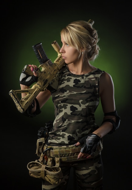 Foto garota com macacão militar de airsoft posando com uma arma nas mãos em um fundo escuro na névoa