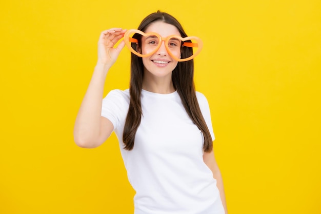 Garota com óculos em forma de coração engraçado e sorrindo no fundo amarelo do estúdio