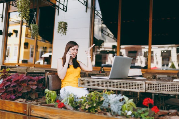 Garota com raiva em um café de rua ao ar livre, sentada à mesa com um computador laptop, falando no celular, gritando e perturbando o problema, no restaurante durante o tempo livre