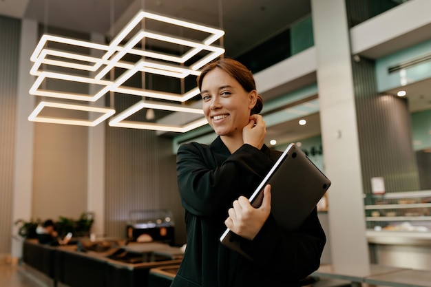 Garota com sorriso terno está de pé no escritório moderno está tocando seu cabelo e olhando para a câmera com um sorriso maravilhoso trabalhadora bonita está se preparando para reunião de negócios