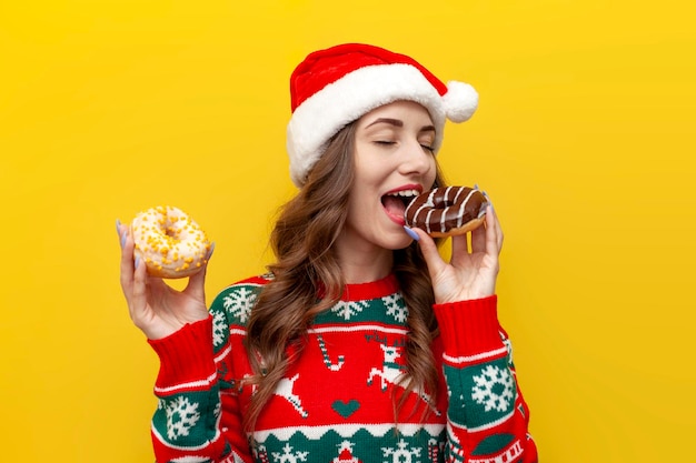 garota com suéter de Natal e chapéu de Papai Noel segura dois donuts e come em fundo amarelo