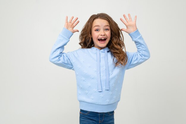 garota com um capuz azul sorri alegremente enquanto segura as palmas das mãos na frente dela em uma parede cinza clara