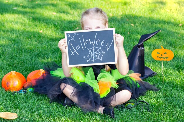 Garota com uma fantasia de bruxa para o feriado de Halloween. Cartaz com a inscrição: Halloween. Garota feliz se senta na grama e fecha os olhos com um cartaz