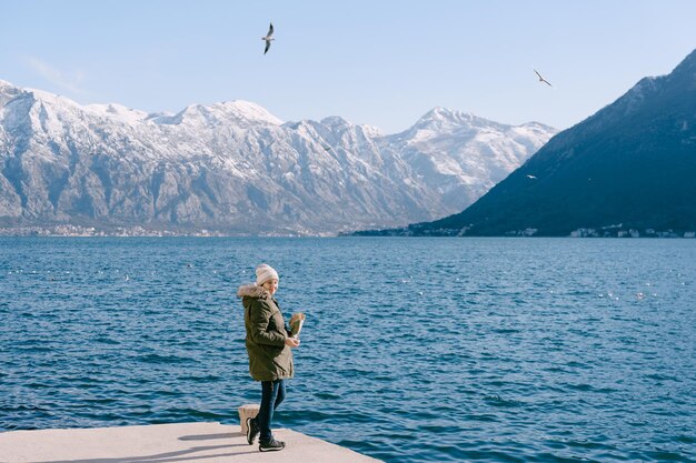 Garota com uma jaqueta fica no cais e alimenta as gaivotas