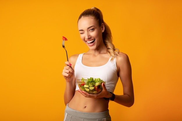 Garota comendo salada piscando para a câmera sobre fundo amarelo