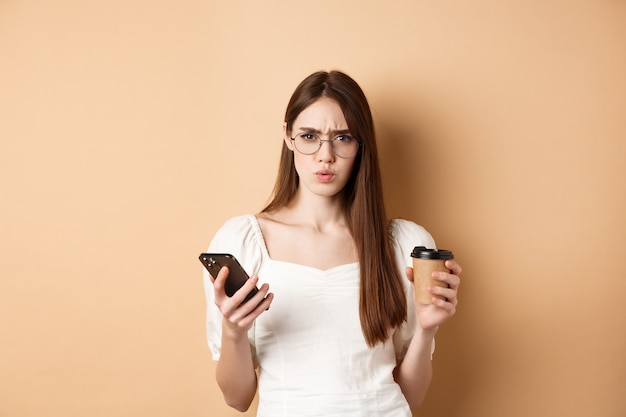 Garota confusa de óculos, franzindo a testa para a câmera depois de ler o telefone móvel, segurando a xícara de café, descontente em pé sobre fundo bege.