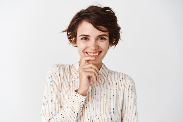 Garota coquete com penteado curto, vestindo blusa, sorrindo romântico e tocando os lábios, feliz, encostada em uma parede branca