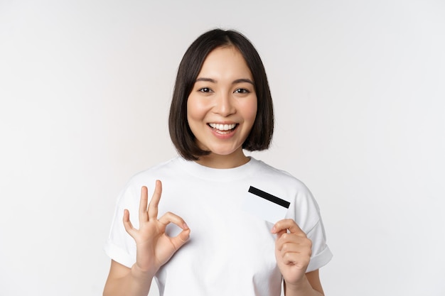 Garota coreana sorridente mostrando sinal de ok e recomendando cartão de crédito do banco de espaço de cópia em camiseta sobre fundo branco
