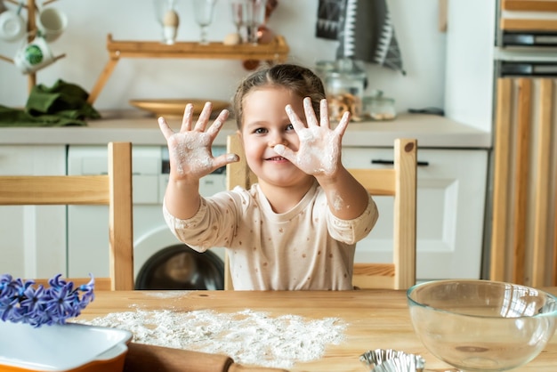 Garota cozinha em casa em uma cozinha brilhante, uma criança agita farinha na mesa, bebê ajuda a mãe e ri