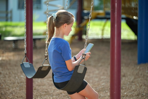 Garota criança feliz navegando em seu celular sentado no balanço no parque durante as férias de verão