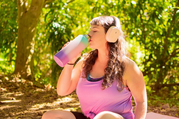 Garota curvilínea com fones de ouvido bebendo água em um parque verde