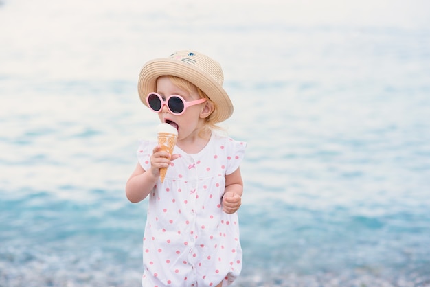 Garota da criança vestida com roupas de verão e óculos de sol rosa fica na praia come com muito prazer sorvete branco. feliz férias de verão.