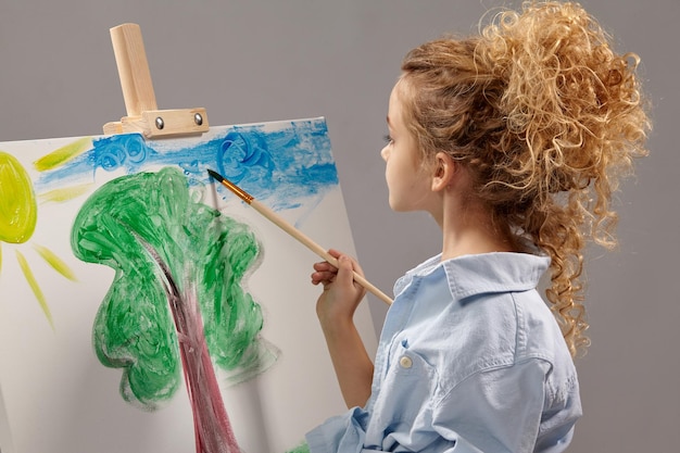 Garota da escola encantadora com cabelo encaracolado, vestindo uma camisa azul e camiseta branca está pintando com um pincel de aquarela em um cavalete, de pé sobre um fundo cinza.