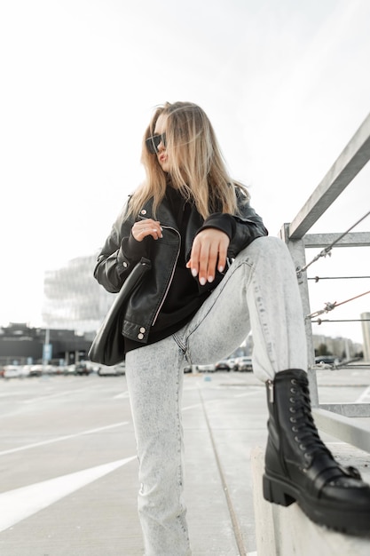 Garota da moda rock legal com jeans de jaqueta preta de couro da moda e sapatos pretos elegantes com poses de bolsa de couro na cidade Moda urbana e beleza