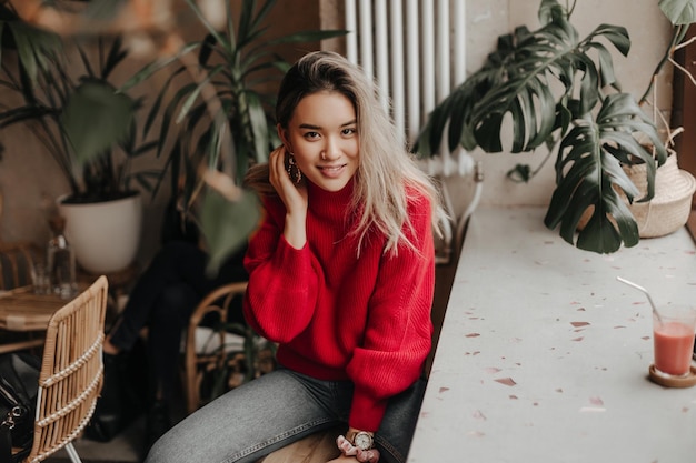 Garota de aparência asiática com cabelo loiro toca suavemente o pescoço e olha para a câmera Retrato de mulher vestida de suéter vermelho e jeans preto sentado no café