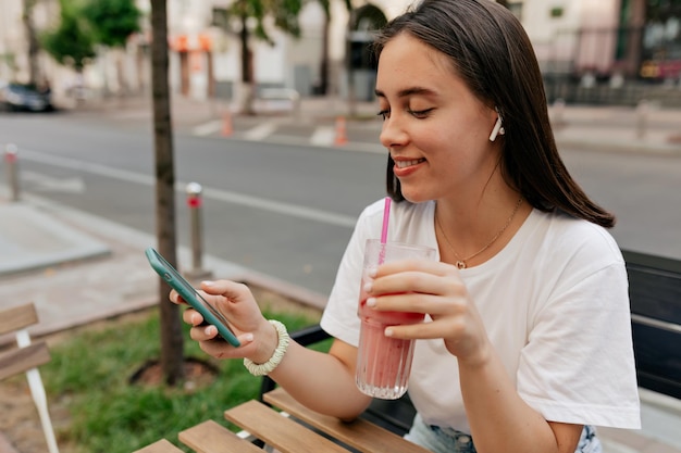 Garota de cabelos escuros animada está rolando smartphone enquanto descansa no café depois de fazer compras Foto ao ar livre de mulher morena feliz está bebendo smoothie