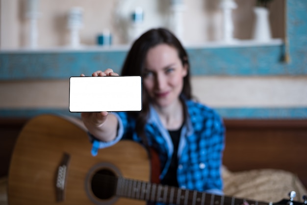 Garota de camisa azul e calça jeans com violão mostra a mão com o smartphone.