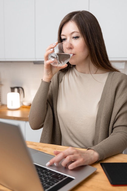 Garota de camiseta bege e jaqueta cinza fez uma pausa no trabalho e bebe água no laptop de mesa