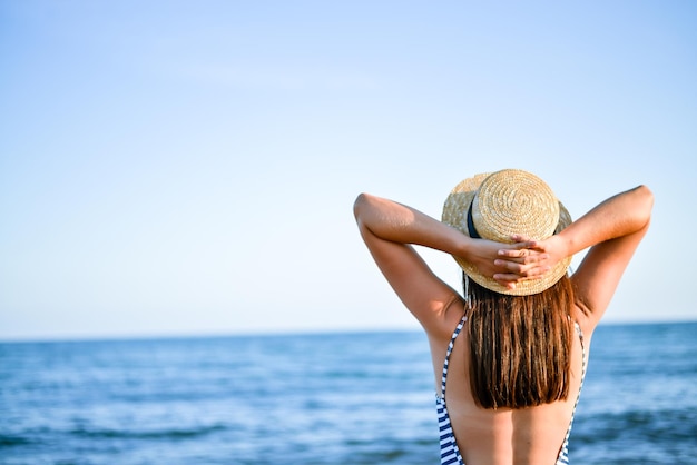 garota de chapéu de palha e maiô listrado, apreciando a vista traseira do mar