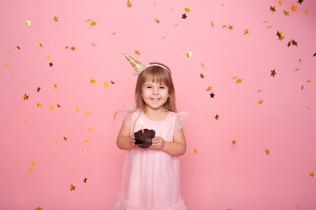 Garota de criança engraçada no vestido com cupcake de aniversário com vela no fundo rosa