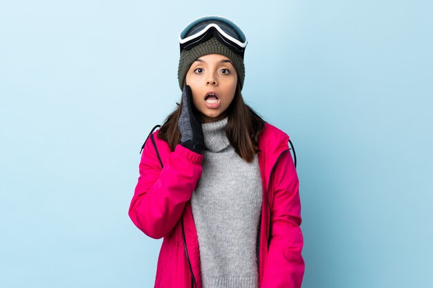 Garota de esquiador de raça mista com óculos de snowboard sobre azul isolado com surpresa e expressão facial chocada.