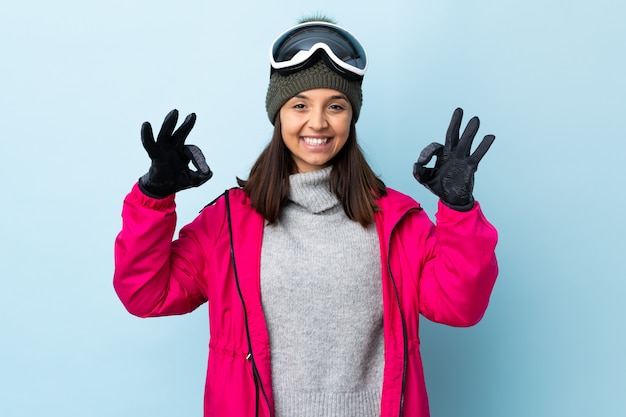 Garota de esquiador de raça mista com óculos de snowboard sobre azul isolado mostrando sinal okey com as duas mãos.