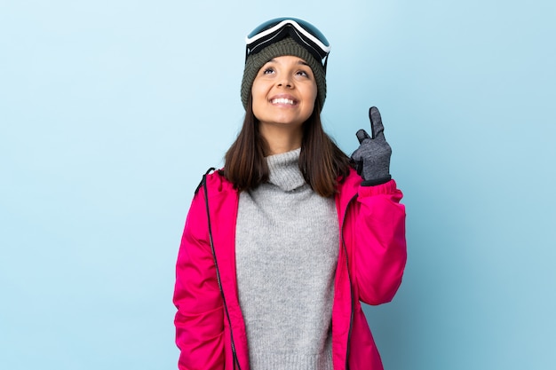 Garota de esquiador de raça mista com óculos de snowboard sobre parede azul apontando para cima e surpreso.