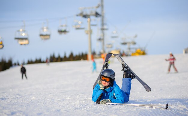 Garota de esquiador deitado na neve em um dia ensolarado contra teleférico na estância de esqui