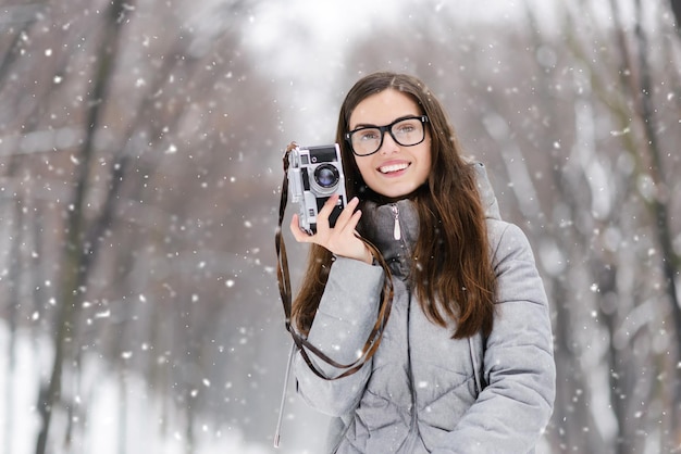 Garota de fotografia morena de óculos vestindo jaqueta cinza segurando câmera retrô na floresta de inverno