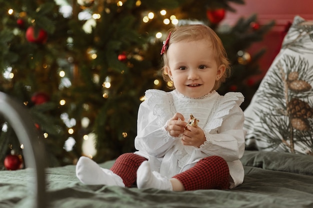 Garota de giro da criança com vestido se senta na cama com luzes de natal festivas ao fundo.