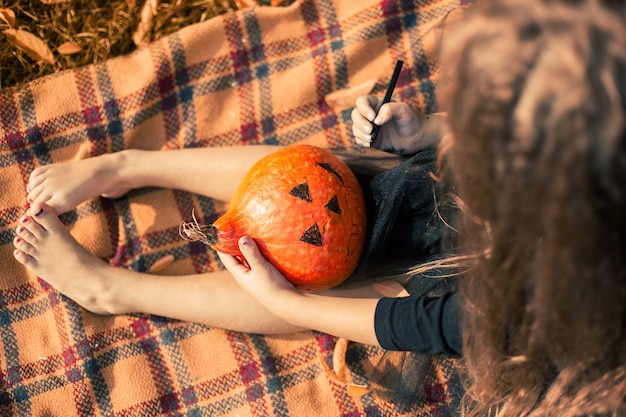 Garota de Halloween com cabelo comprido, vestido preto senta-se em um cobertor laranja no parque e desenha um ...