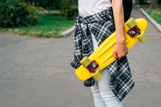 Garota de jeans e uma camisa xadrez está andando no parque com skate de plástico amarelo nas mãos