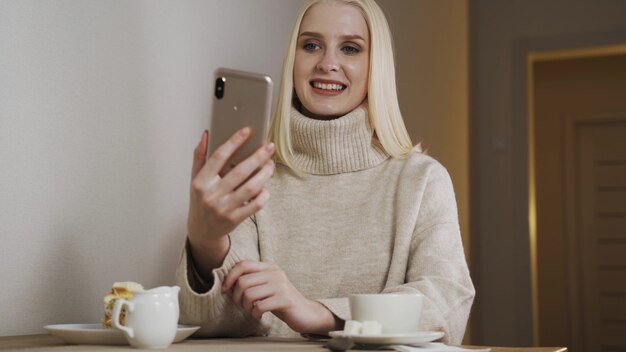 Garota de jovem estudante atraente muito feminino usando telefone celular, bebendo café no café à noite, passar o tempo online rindo mensagens de bate-papo.