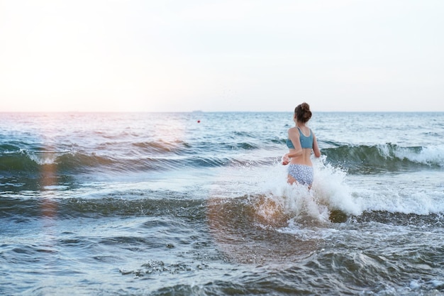 Garota de maiô está brincando na praia com uma onda do mar pulando correndo se divertindo