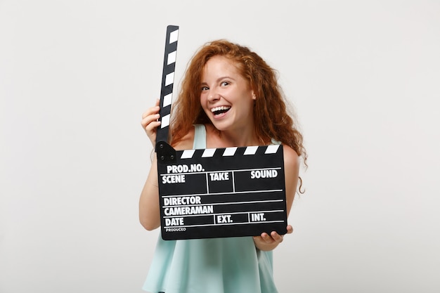 Garota de mulher jovem ruiva engraçada em roupas leves casuais posando isolado no fundo da parede branca no estúdio. Conceito de estilo de vida de pessoas. Simule o espaço da cópia. Segurando o clássico filme preto fazendo claquete.