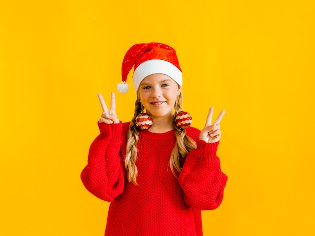 Garota de Natal. Linda garotinha com um chapéu de Papai Noel vermelho mostra legal. Garota com uma fantasia de ano novo em um fundo amarelo.