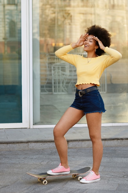 Garota de pele escura em um top amarelo e shorts azul com um skate contra o espaço do verão de parede na cidade