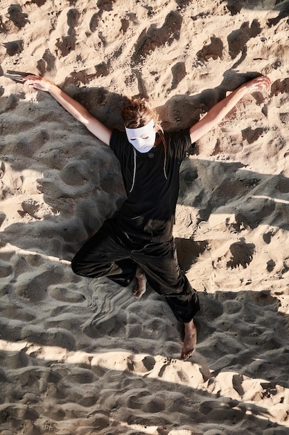 Garota de performance teatral com máscara de teatro branca encontra-se na areia do mar Performance de teatro ao ar livre da dançarina em dia ensolarado Mulher de máscara branca e estilo preto total de pano na vista superior da costa do mar