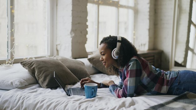 Garota de raça mista atraente ouve música enquanto navega nas mídias sociais no laptop deitada na cama