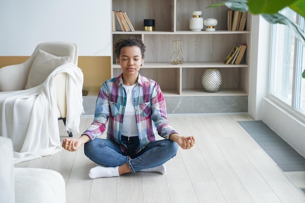 Garota de raça mista fazendo ioga medita para aliviar o estresse sentado em pose de lótus no chão em casa