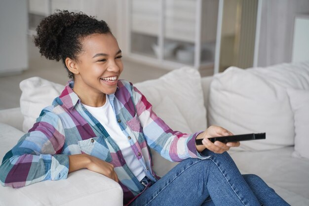 Garota de raça mista rindo assistindo TV comédia show filme série de televisão sentada no sofá em casa