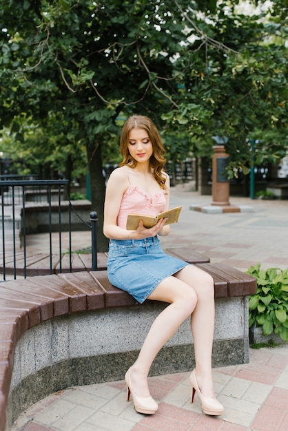 garota de saia rosa top e jeans, lendo um livro enquanto está sentado em um banco