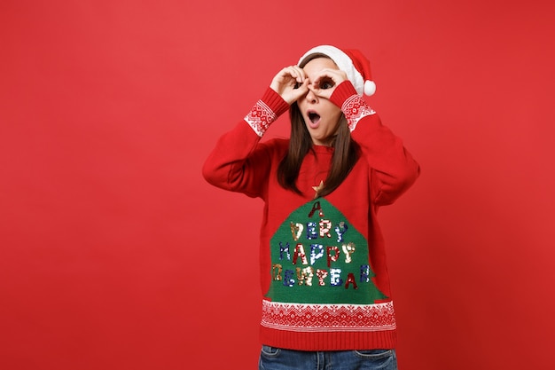 Garota de Santa surpresa, mantendo a boca aberta, de mãos dadas perto dos olhos, imitando óculos ou binóculos isolados sobre fundo vermelho. Conceito de festa de feriado de celebração de ano novo feliz 2019. Simule o espaço da cópia.