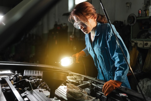 Garota de serralheiro automático inspeciona o motor do carro iluminando a luz da lâmpada Garagem ou oficina e uma mulher no trabalho de macacão e óculos