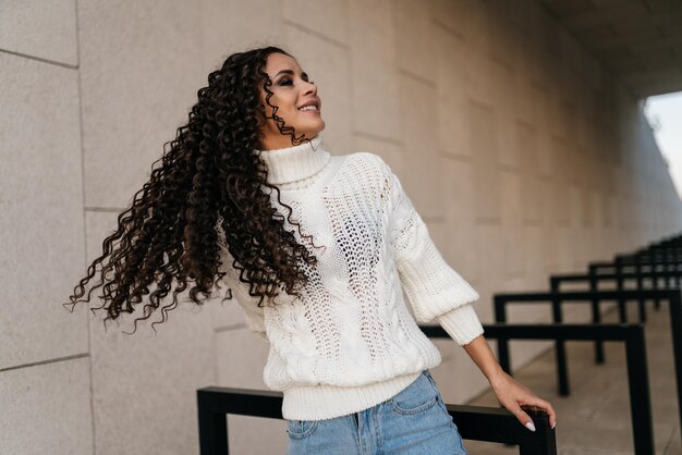 Garota de suéter branco e jeans torce a cabeça jogando cabelo longo encaracolado