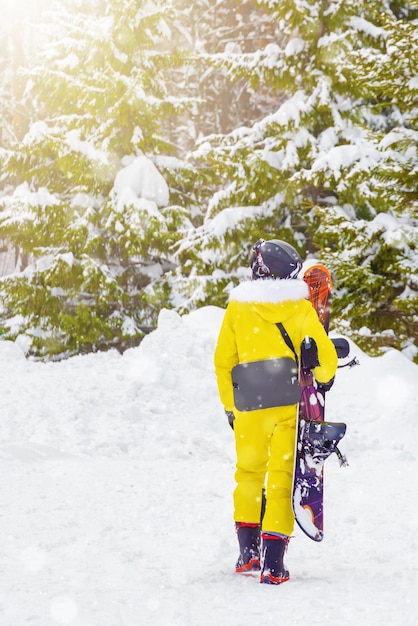 Garota de terno amarelo de snowboard com snowboard nas mãos no fundo da floresta de inverno vai passear
