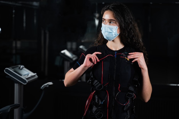 Garota de terno EMS e máscara médica no ginásio. Proteção contra coronavírus covid-19. Treinamento esportivo em traje de estimulação elétrica muscular.
